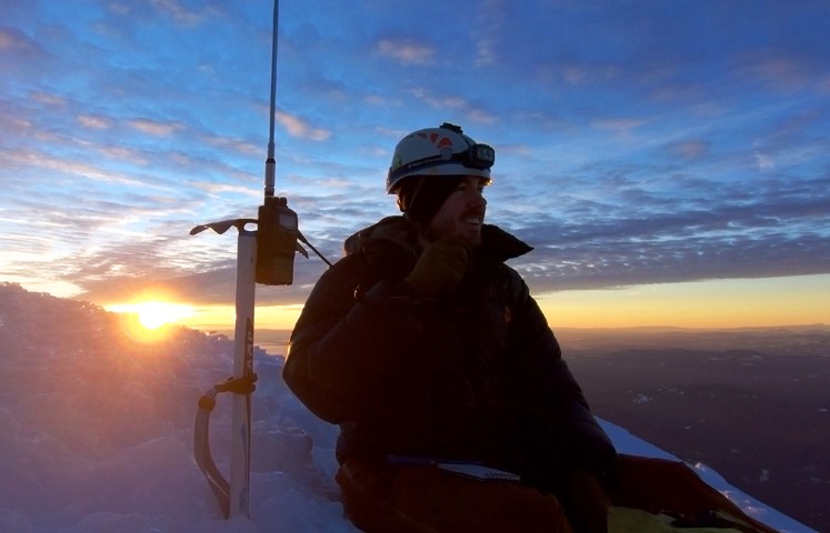Tim in a black puffy jacket sitting on the top of Mt Hood. The sunrise is behind him. A handheld radio is clipped to an ice axe standing in the snow.